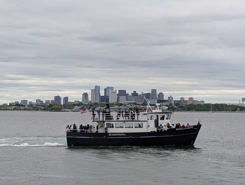 The image shows a black and white boat sailing on the water. The boat is filled with people and has the name "Beverly Ann" on its side. In the background, there is a city skyline under a cloudy sky.
